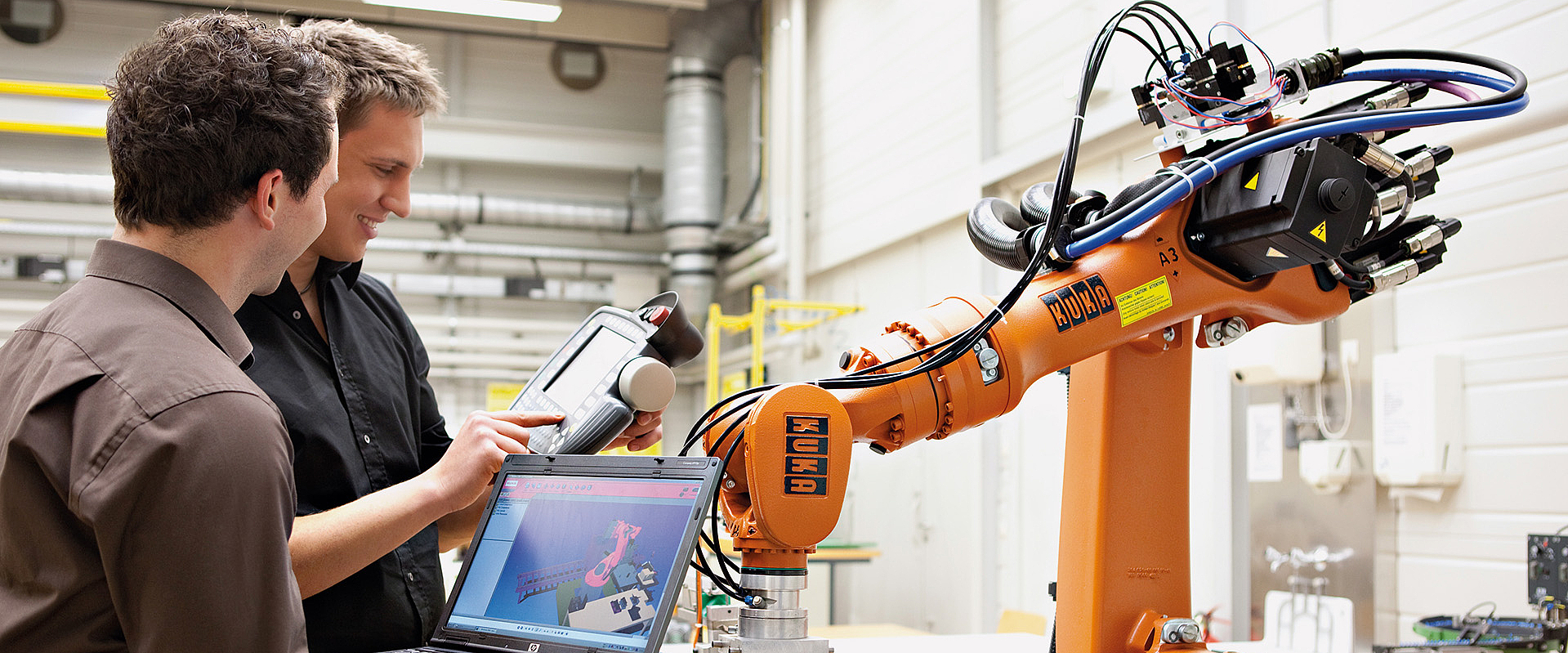 Zwei Maschinenbaustudierende stehen mit Tablet vor einem orangenen KUKA-Roboter