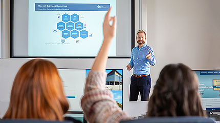 Professor steht vorne mit Präsentation zu digitalem Marketing und ruft freundlich Studentin auf