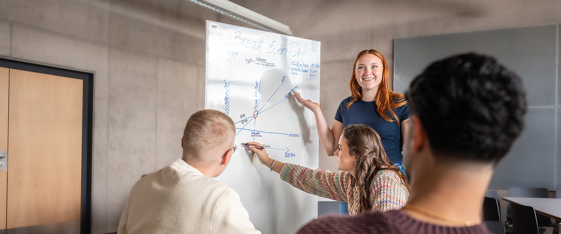 Studentengruppe arbeitet an Whiteboardtafel. Rothaarige Studentin steht am Whiteboard und deutet mit dem Stift auf die Tafel. Sie spricht zu Ihren Kommilitonen