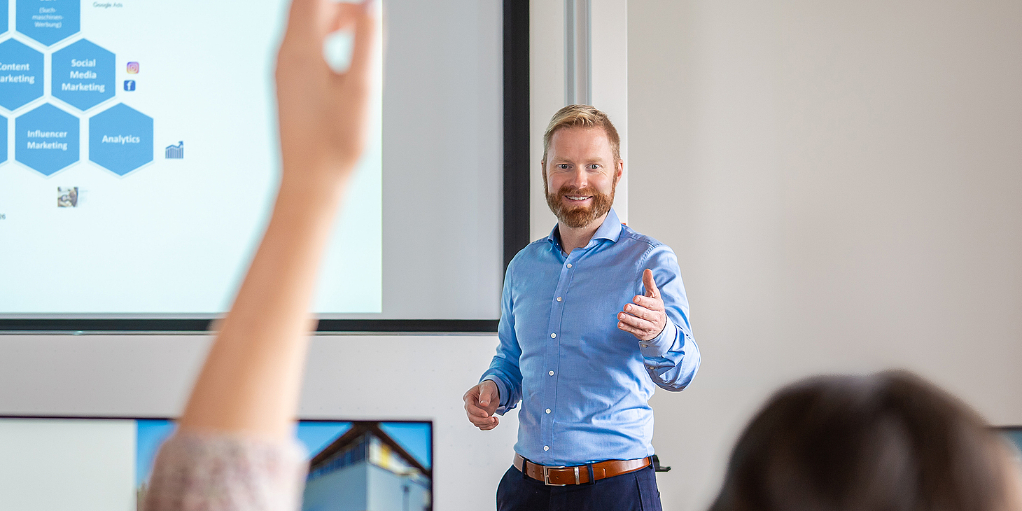 Prof. Dr. Martin Haupt steht vor einer Projektion seiner Präsentation und ruft freundlich eine Studentin auf