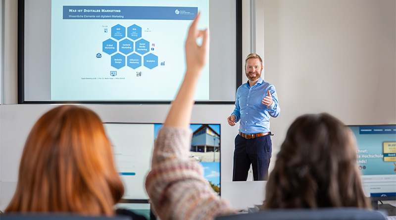 Professor Dr. Martin Haupt Ruft Studentin in der Vorlesung auf. Zu sehen ist eine Präsentation an der Wand. Die Studentin ist von hinten, der Professor frontal zu sehen